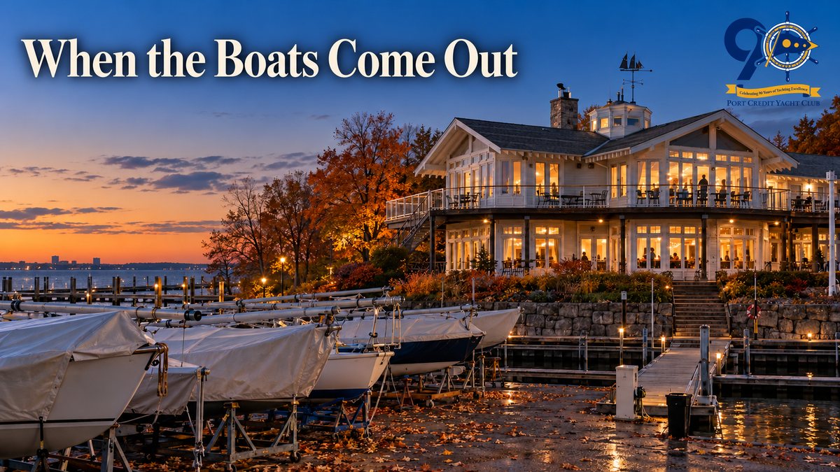 Sailboats on cradles under covers in the Port Credit Yacht Club yard on an autumn evening, masts down and fallen leaves on the ground, with the empty docks below and the clubhouse windows lit warm against a deep blue dusk sky.